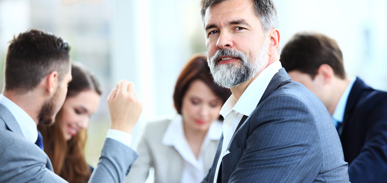 Un groupe de gens d’affaires assis autour d’une table en train de discuter, avec un homme tourné vers l’arrière souriant à la caméra