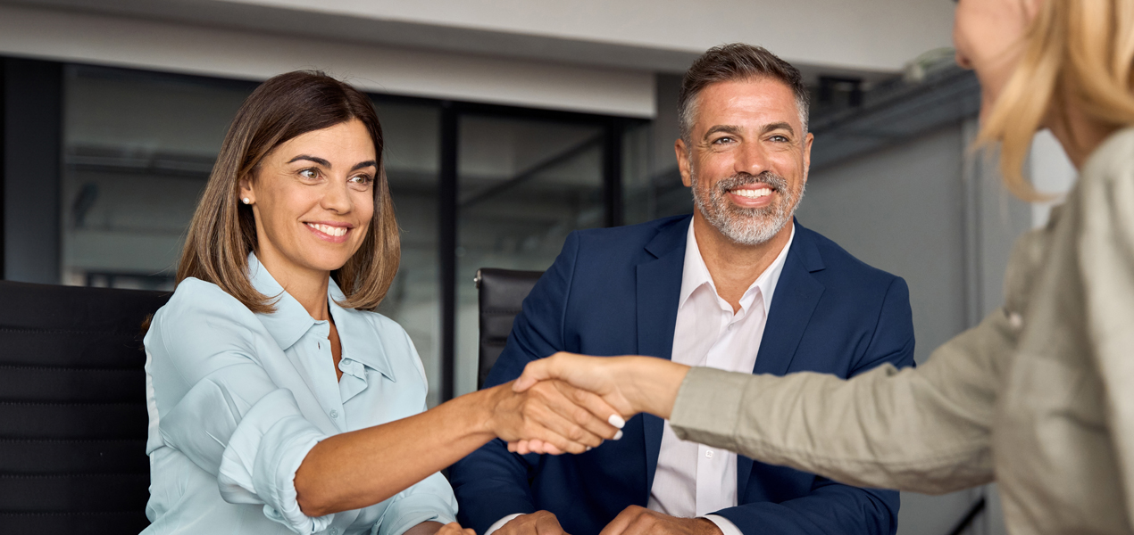 Deux professionnels, une femme en chemisier bleu clair et un homme en costume bleu, sourient en serrant la main d'une personne au premier plan. Ils sont assis dans un cadre de bureau