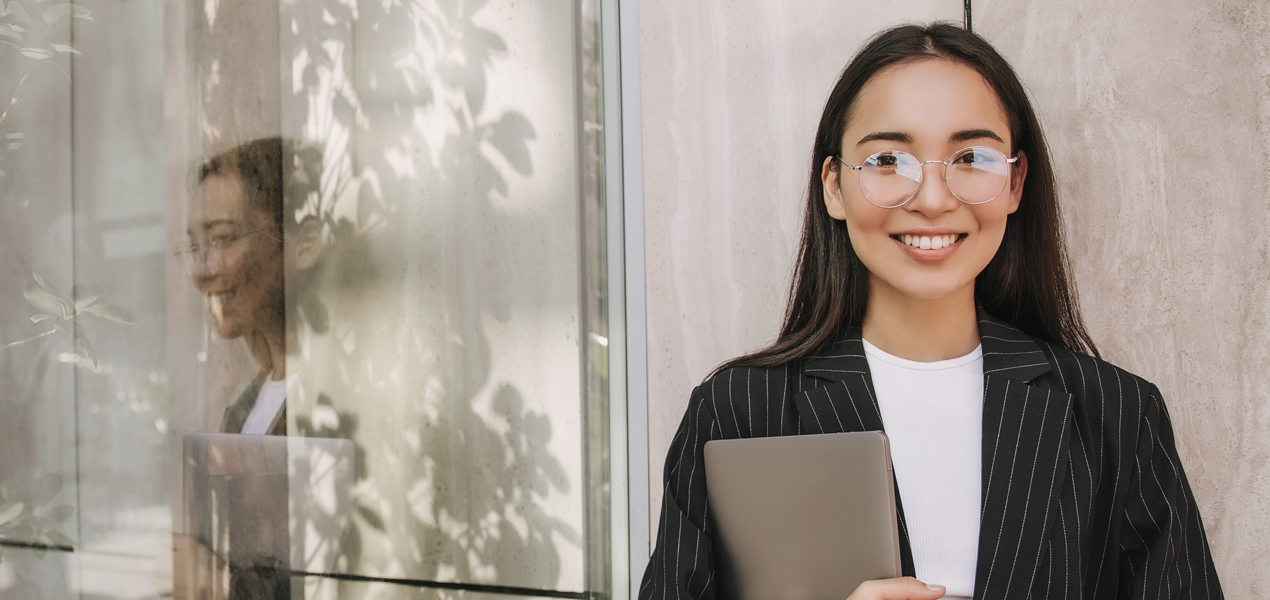 Étudiante à côté d’une fenêtre, tenant son ordinateur portable et souriant à la caméra.