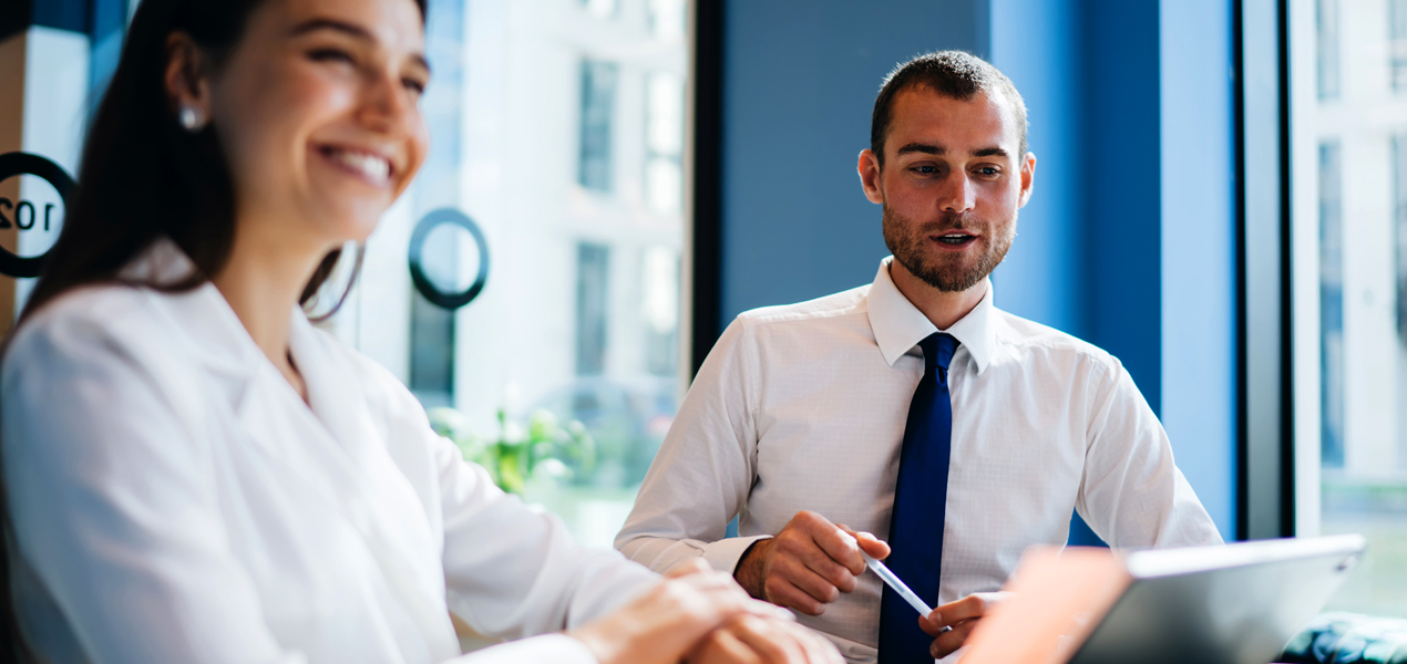 Deux personnes en tenue professionnelle sont assises à une table dans un bureau moderne aux grandes fenêtres. L’une sourit, tandis que l’autre est concentrée sur une tablette, un stylo à la main. Des 
