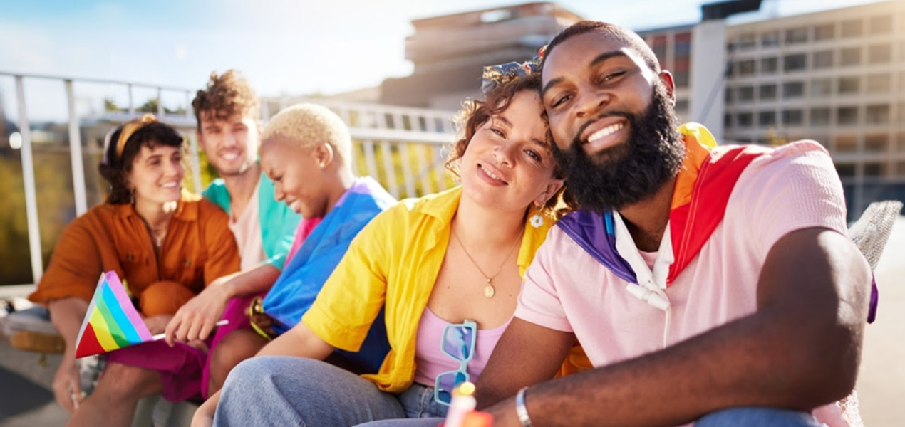 Un groupe diversifié de personnes tenant des drapeaux arc-en-ciel, assises ensemble sur un banc, célébrant la fierté et l’inclusion.