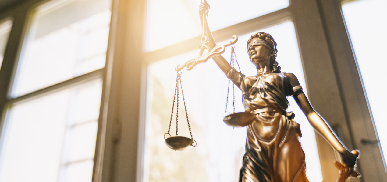 Statue en bronze de la Justice tenant une balance et une épée, placée devant une grande fenêtre baignée de lumière, symbolisant l’équité et la loi. La figure est les yeux bandés